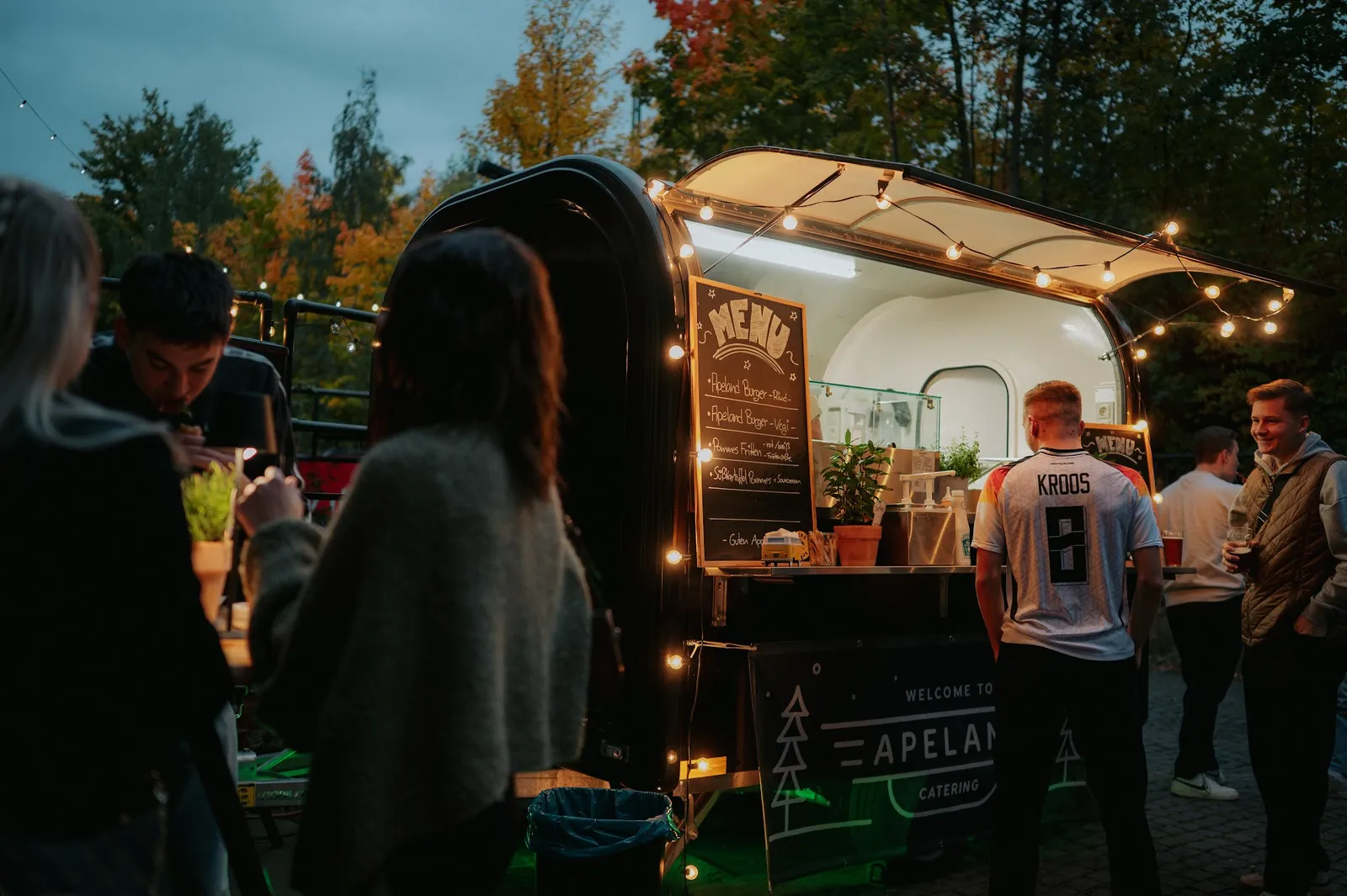 Gäste stehen abends entspannt vor dem beleuchteten Foodtrailer